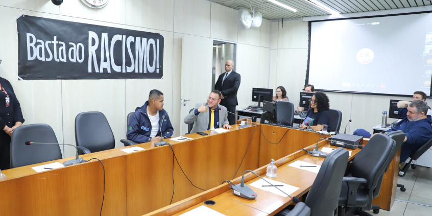 Imagem dos participantes da Audiência Pública. Uma faixa negra pendurada na parede traz os dizeres: Basta ao racismo