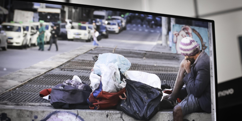 Pessoa em situação de rua sentada na calaçada diante de seus pertences. O homem usa uma blusa de brio, touca e bermuda