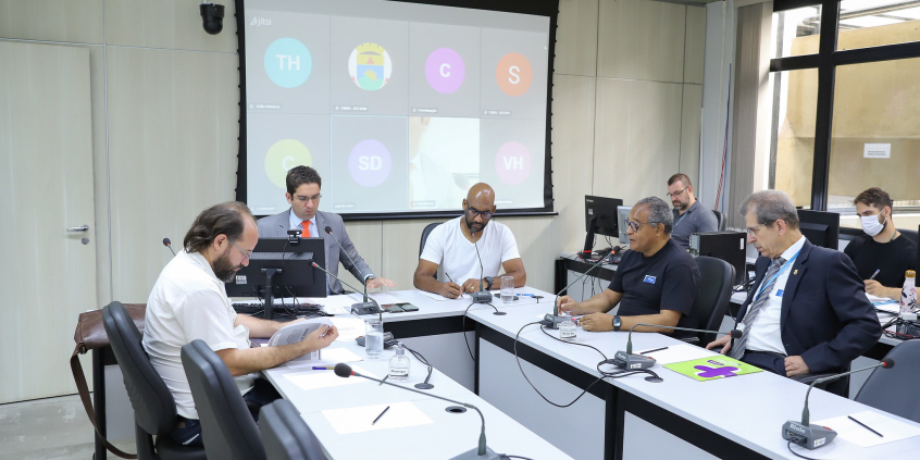 Cinco homens sentados à mesa, em reunião presencial.