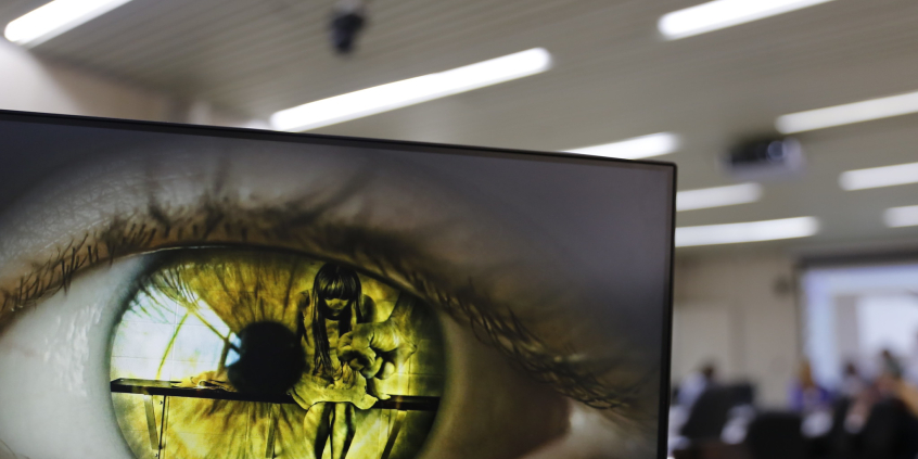 Imagem de um olho verde na tela do computado. É possível identificar uma menina de cabeça baixa refletida na imagem