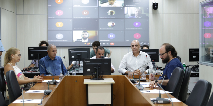 Quatro parlamentares e uma representante sindical reunidos, sentados à mesa. Ao fundo, telão exibe participação remota de representantes da Secretaria Municipal de Saúde.