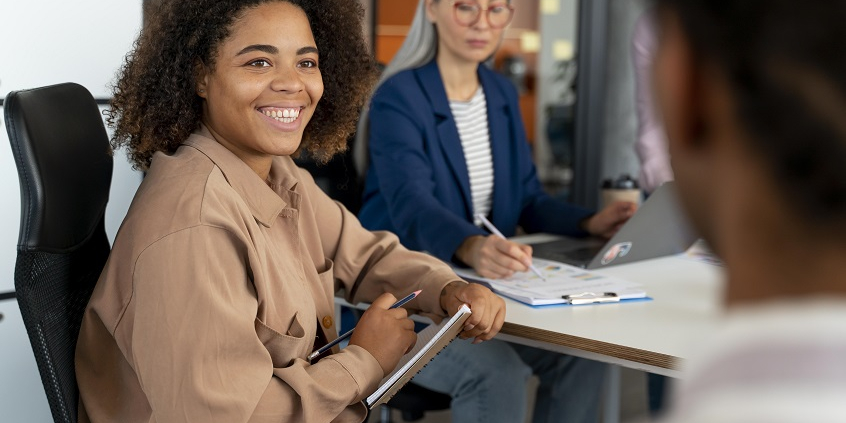 Mulher negra sentada em uma cadeira faz anotações em caderno. Atrás dela está uma mulher mais velha de cabelos grisalhos longos, também fazendo anotações. À frente dela, de costas e desfocada, está uma pessoa em pé, ensinando.