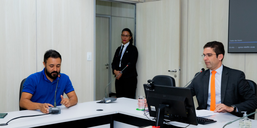 Os vereadores Braulio Lara e José Ferreira estão sentados no plenário. Uma segurança está em pé ao fundo, junto à porta.