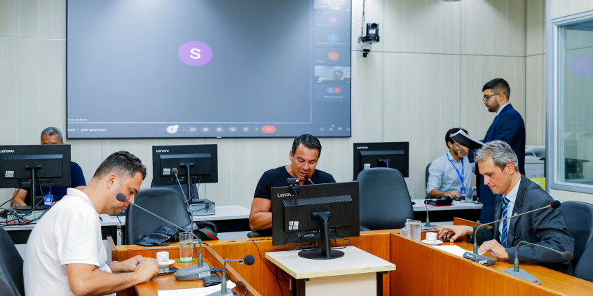 Vereadores discutem projetos no Plenário Camil Caram. O telão atrás deles mostra videochamada com vereadores presentes virtualmente.