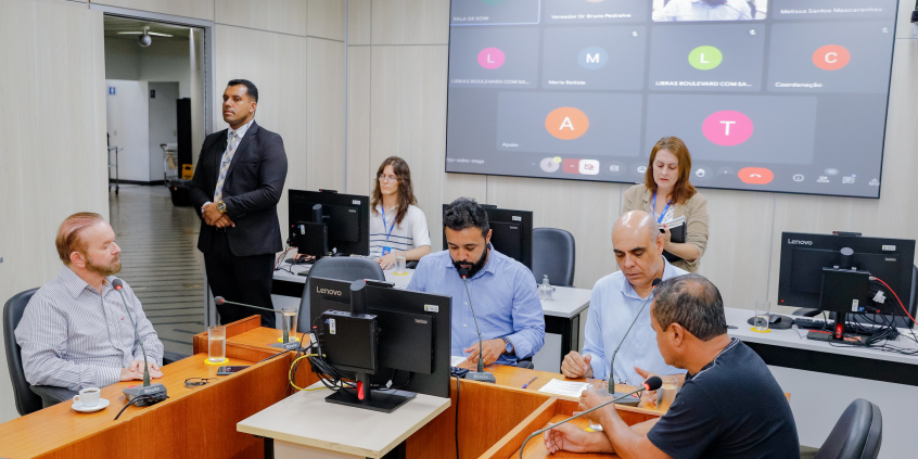 Vereadores estão sentados no plenário Camil Caram. O telão atrás deles mostra a videochamada com um parlamentar presente virtualmente.