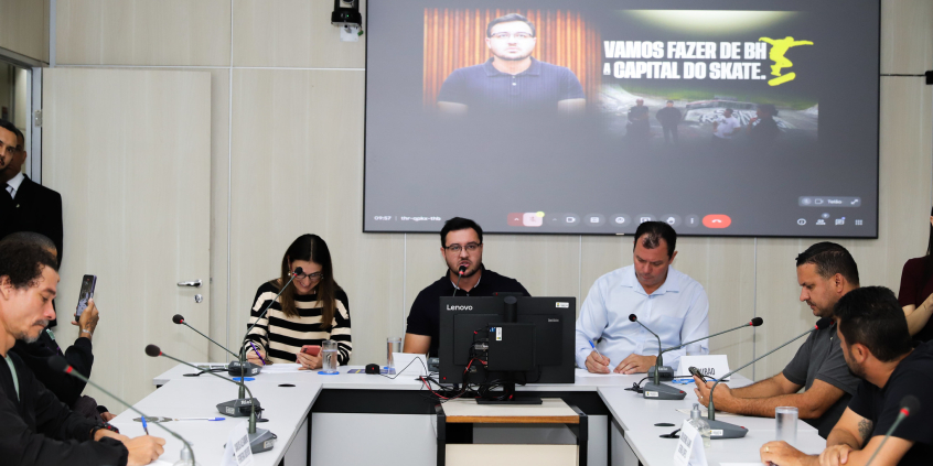 Vereadores e convidados assentados no Plenário Helvécio Arantes durante audiência pública. O telão atrás deles mostra apresentação.