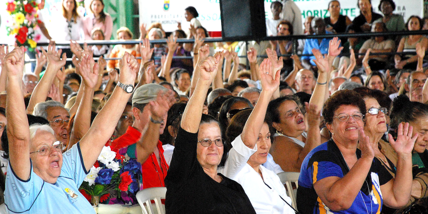 pessoas idosas assentadas levantam os braços participando de evento