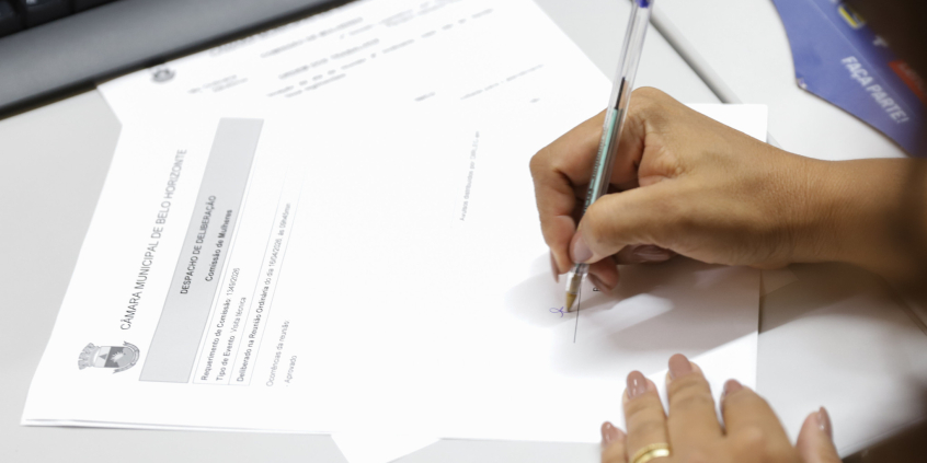Mão de vereadora assina documento durante reunião da Comissão de Mulheres