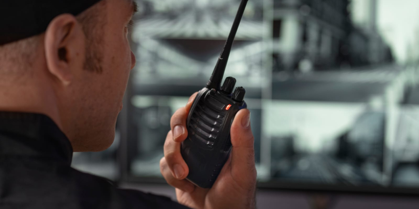 Vigilante aparece de lado, falando em walkie talkie