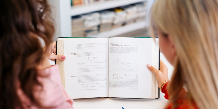 Duas mulheres de costas leem livro didático