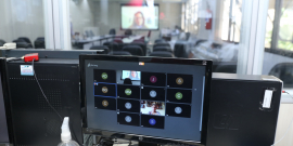 Tela de computador exibindo videoconferencia. Ao fundo, cadeiras vazias no plenário e telão exibindo videoconferência