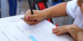 Mãos de criança escrevendo em papel sobre a mesa. Papel exibe atividade escolar