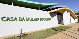 Fachada da Casa da Mulher Brasileira em Brasília. Muro branco com o nome da unidade em verde. Teto ondulado nas cores verde, azul e amarela