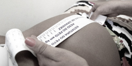 Barriga de mulher grávida em detalhe. Mãos de outra mulher faz a medição do crescimento do bebê, em acompanhamento pré-natal