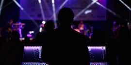 Em boate com luz azulada, DJ, de costas, atrás de uma mesa de som, observa pessoas dançando no fundo. 