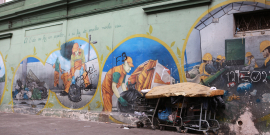 Em frente a um muro pintado com imagens de homens trabalhando, há trêsx carrinhos de supermercado cobertos com um colchão, indicando que há um pessoa habitando o local.
