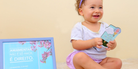 Criança caucasiana usa lado de fita lilás no cabelo, blusa branca e short lilás. ao lado, um quadro com os dizeres: "Amamentar não é só amor. É um direito."