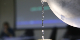 Um jarro de água despeja o líquido em um copo transparente . Em segundo plano, a tela do computador com imagem desfocada