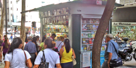 Várias pessoas passam diante de uma banca de revista no centro da cidade