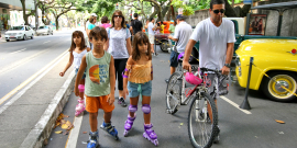 Crianças brincam na rua com patins ao lado de um adulto que está de bicicleta