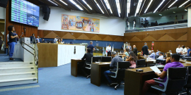 Imagem lateral do Plenário Amynthas de Barros. Estão enquadrados a mesa diretora e as cadeiras dos parlamentares. Ao fundo e no alto, o painel de Yara Tupinambá