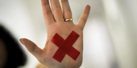 foto mostra uma mão aberta, com a palma virada para frente, uma aliança no dedo anelar e um "X" vermelho colado na palma.