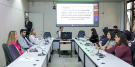 Osvaldo Lopes e convidados da audiência pública assentados no Plenário Helvécio Arantes, tela atrás deles mostra apresentação sendo realizada