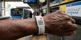 Braço de um homem mostrando pulseira branca de silicone com QR Code. Ao fundo existe uma estação de metrô de Belo Horizonte. Imagem criada com auxílio de Inteligência Artificial