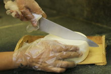 Queijo branco em tábua de madeira sendo cortado por mãos enluvadas.