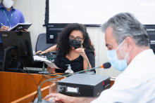 Dois vereadores sentados a mesa participando de reunião, em uma sala. 