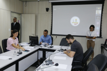 Três membros da Comissão em reunião, em torno de uma mesa em formato de U. Na imagem é possível ver um segurança na porta e uma servidora de pé auxiliando na reunião