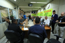 Sala de reuniões com muitos homens e mulheres sentados e de pé, cartazes com dizeres como "Caçamba não é lixo" não paredes. 