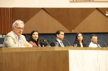 TRês homens e duas mulheres sentados à mesa. O primeiro deles, à esquerda, palestra.