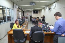 Dois parlamentares reunidos, sentados à mesa, auxiliados por um assessor técnico da câmara, de pé, ao lado deles. ao fundo, mais de dez pessoas assistem à reunião, sentadas. 