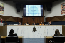 Três mulheres e dois homens sentados à mesa, com alguns expectadores à frente.