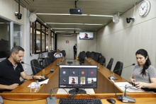 dois parlamentares sentados à mesa, em reunião presencial, e dois participando remotamente, exibidos em tela de computador.ticipando 