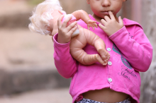 Imagem de uma criança pequena com dedo na boca, usando blusa rosa e carregando uma boneca. 