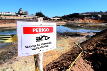 Foto de barragem ao fundo com placa a frente escrito "serviço de escavação valas abertas"