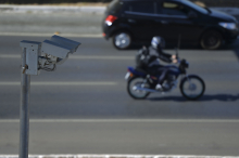 Foto mostra, em primeiro plano, câmera de monitoramento e, na rua, moto e automóvel