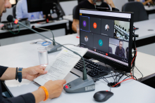 Tela do computador mostra reunião virtual dos vereadores. Em frente à tela, duas mãos seguram um documento com a pauta da reunião.